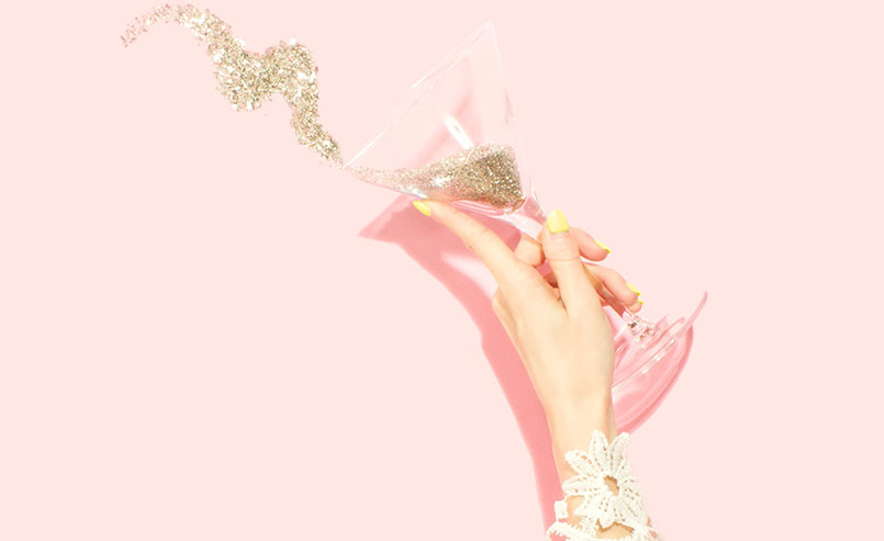 Mão feminina, com unhas amarelas e traje rendado segurando uma taça de martini com glitter em fundo rosa.