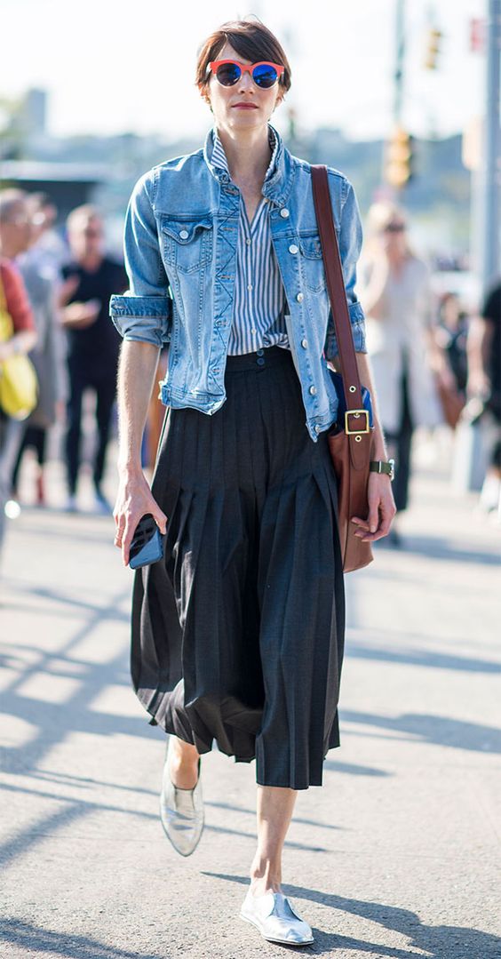 mulher morena e magra veste camisa de listras, jaqueta jeans com as mangas arregaçadas, saia plissada midi e oxford metalizado